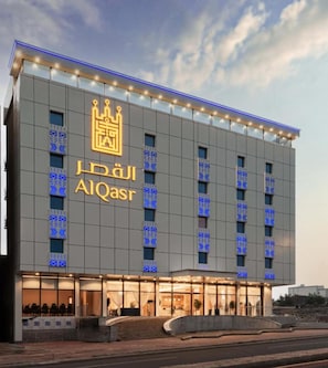 A modern hotel building with a prominent crown logo and the name 'AIQasr' displayed on the facade.
