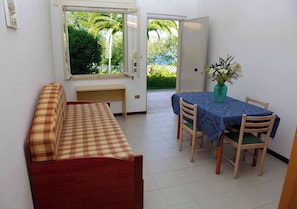 A room with a bed, a dining table with chairs, a window with a view of greenery, and a door.