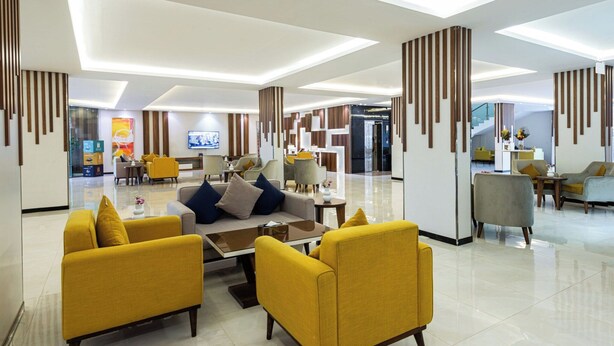 Um saguão de hotel moderno, com área de estar central, balcão de recepção e escadaria para os outros andares.