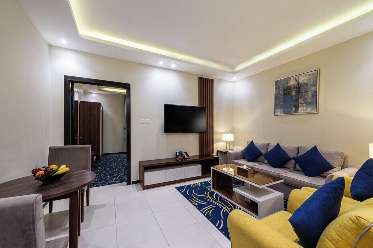 A modern hotel room with a flat-screen TV, a sofa with blue cushions, a dining table with fruit, and a patterned rug.