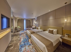 A hotel room with two beds, a desk, a chair, a TV, and a large window with curtains. - Grand Continent Luxury Collection, Udaipur (Udaipur)