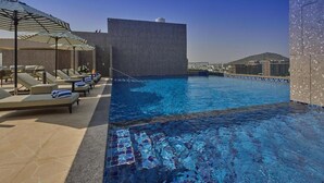 A rooftop pool with lounge chairs and umbrellas, a clear sky, and a distant cityscape. - Grand Continent Luxury Collection, Udaipur (Udaipur)