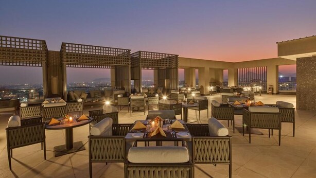 An outdoor dining area with tables and chairs, a fire feature, and a city view at dusk. - Grand Continent Luxury Collection, Udaipur (Udaipur)