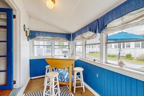 A room with a wooden table, two white barstools, a striped rug, and a window with a view of a blue umbrella and a fence.
