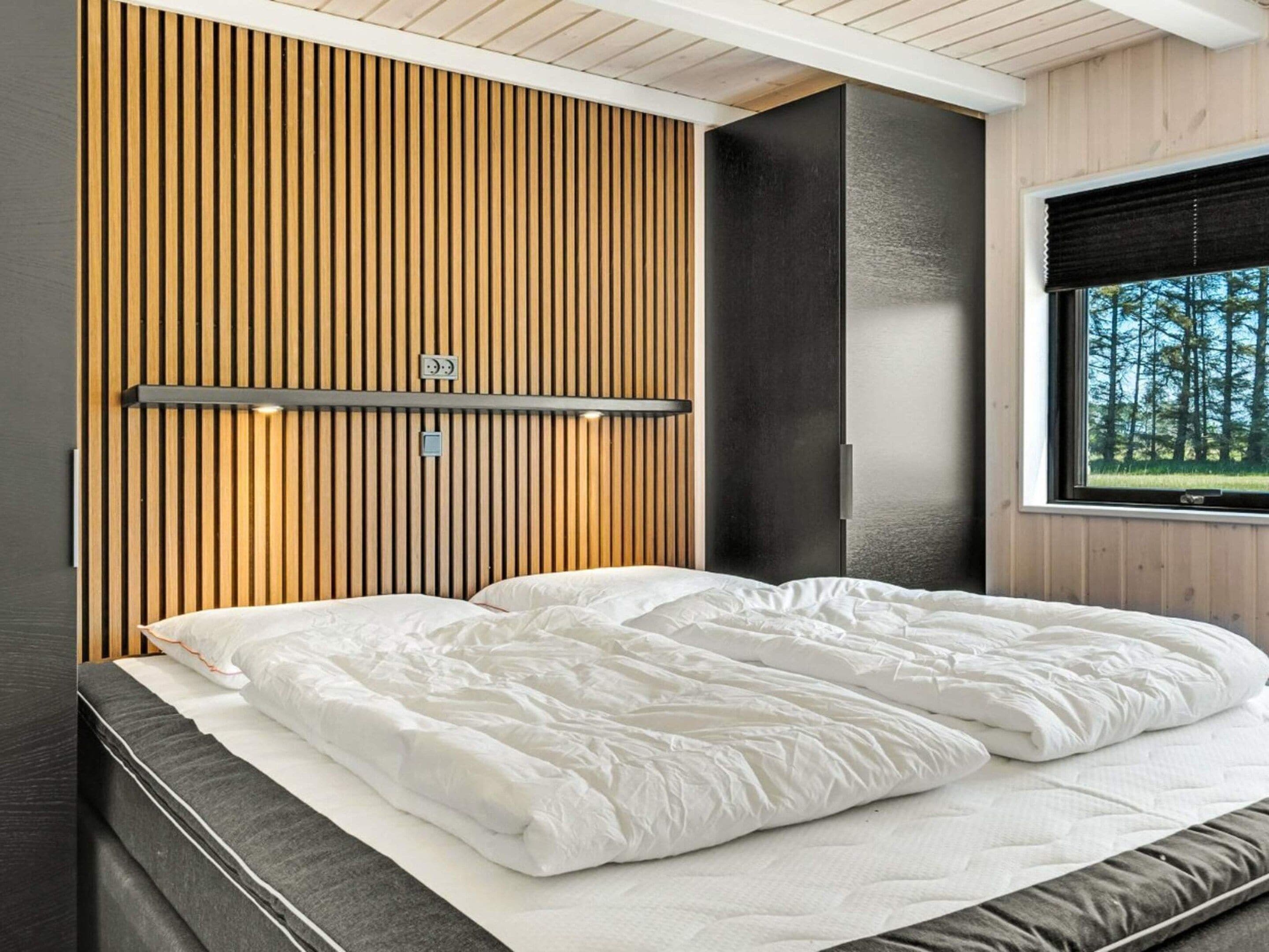 Un dormitorio moderno con cama, panel de pared de madera y ventana con vistas a árboles.