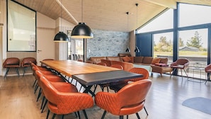 Un vaste espace dédié à la salle à manger, avec une longue table en bois, de nombreuses chaises orange et des lampes suspendues noires.