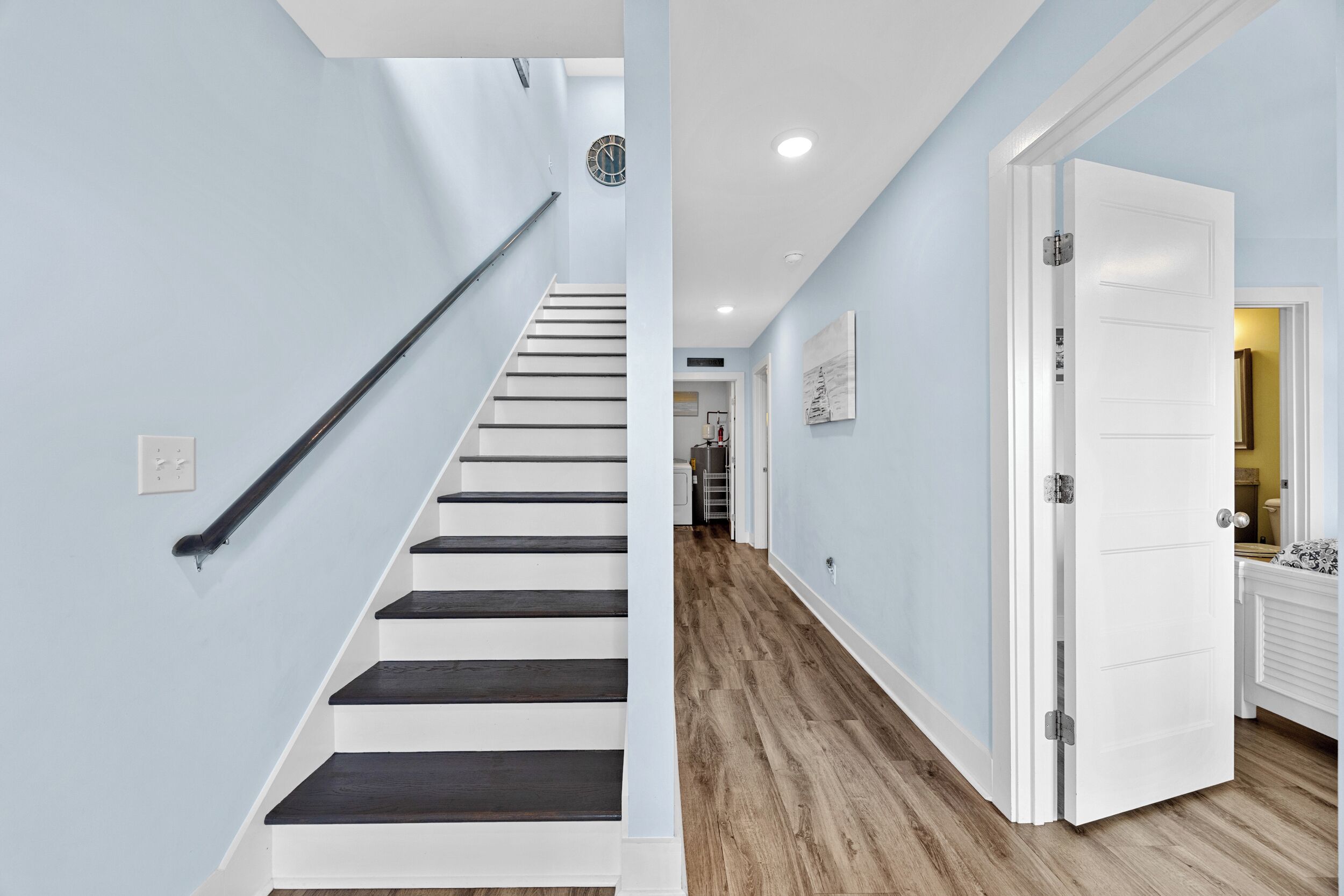 Un escalier aux murs blancs et aux marches sombres, une porte blanche avec une poignée argentée et un couloir au plancher en bois.