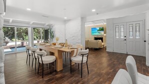 Een ruime eethoek met een houten tafel en witte stoelen, een witte bakstenen muur, en een groot raam met uitzicht op groen.