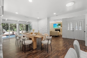 A spacious dining area with a wooden table and white chairs, a white brick wall, and a large window with a view of greenery.