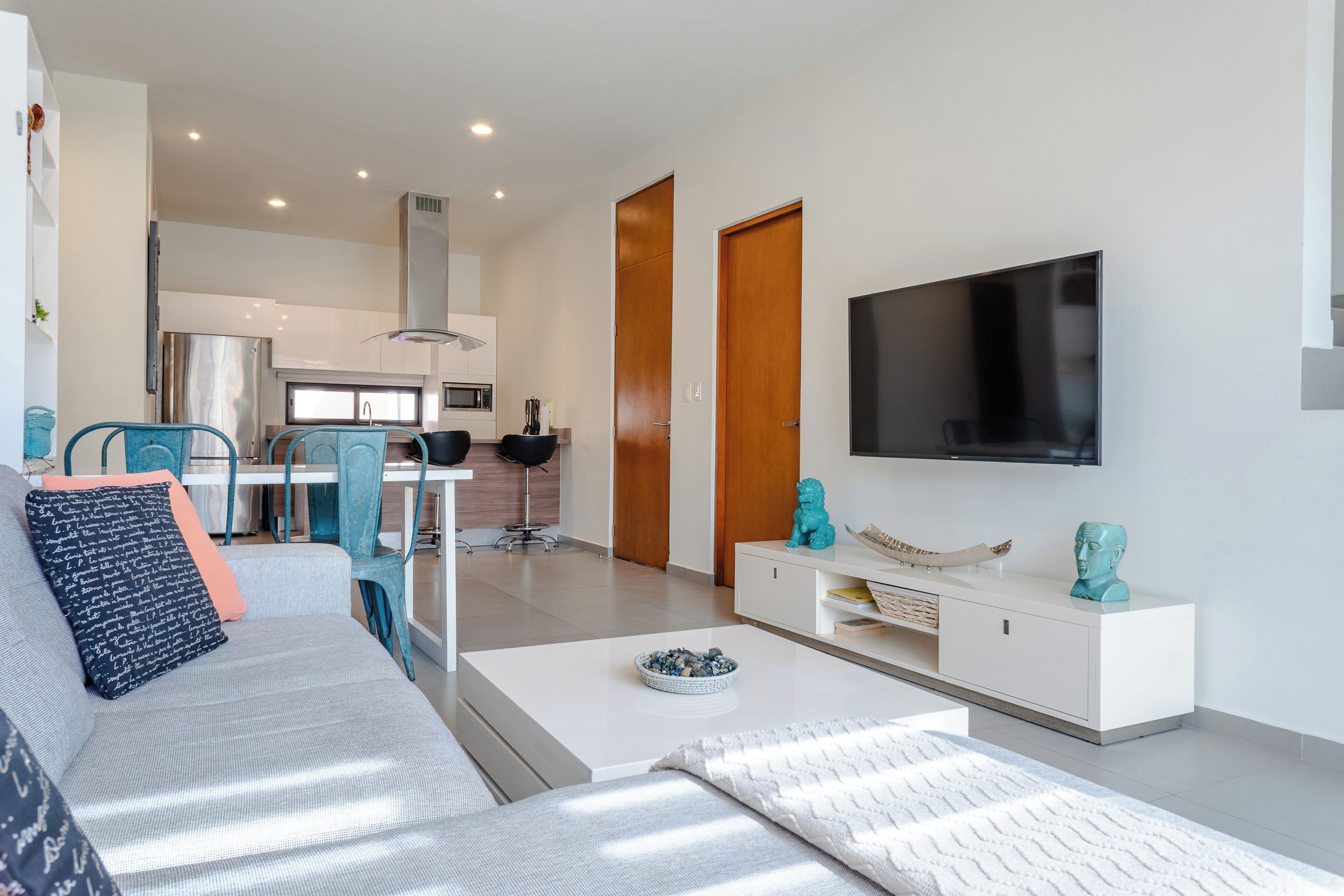 Sala moderna com sofá, mesa de centro e televisão. Cozinha visível ao fundo, com armários e micro-ondas.