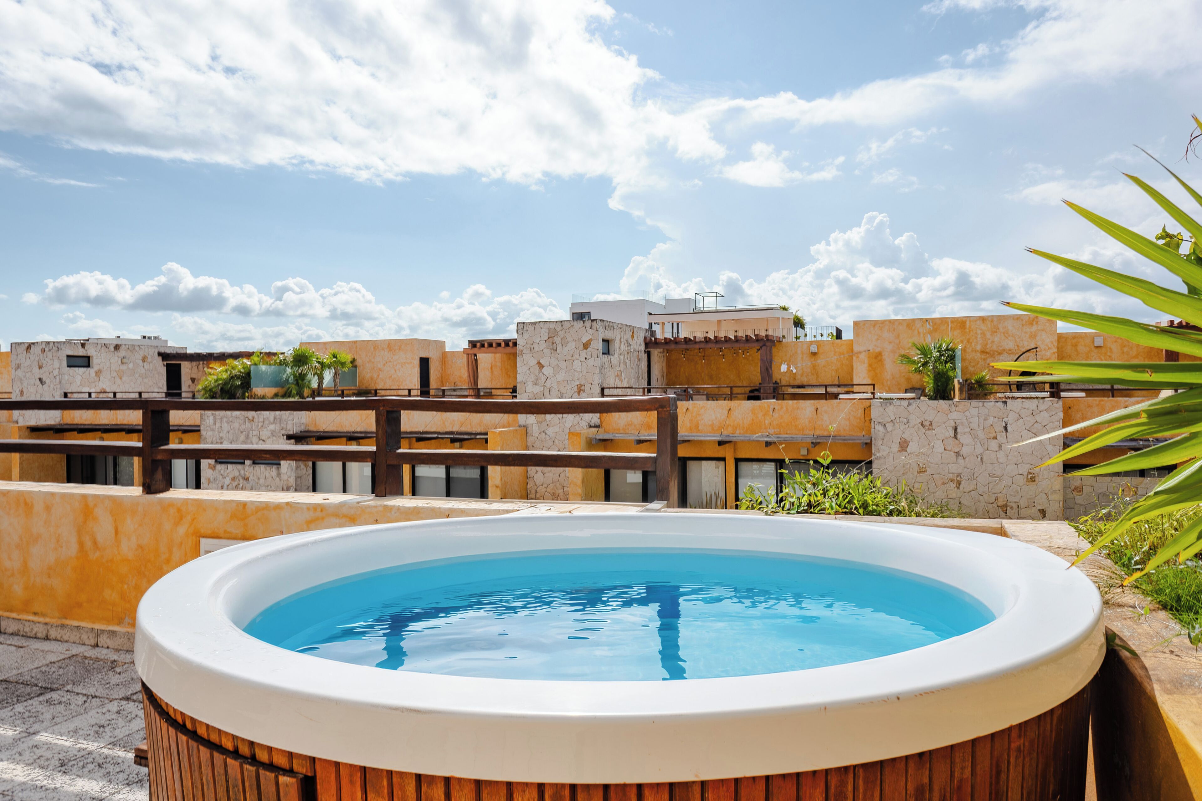 A rooftop swimming pool with a wooden deck, surrounded by buildings with a stone facade and a clear sky.