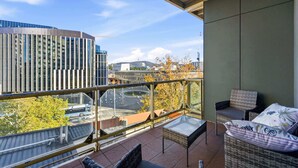Un balcone con ringhiera in vetro, mobili in vimini e vista sugli edifici della città e su una strada.