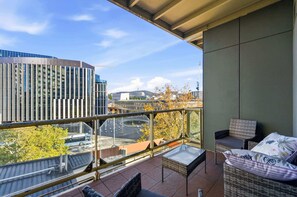 Ein Balkon mit Glasgeländer, Korbmöbeln und Blick auf Stadtgebäude und eine Straße.