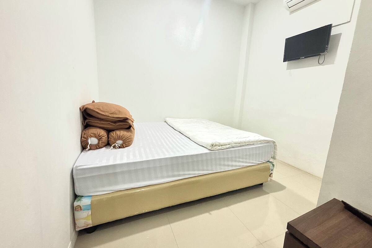 A small, single-bed room with a white mattress and a beige headboard. There is a brown pillow on the bed, and a small brown table is visible in the corner.