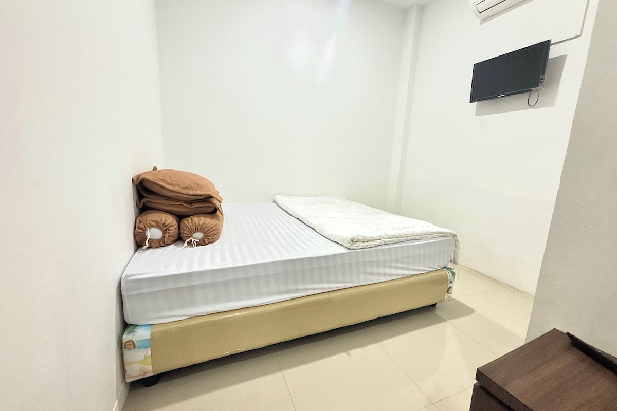 A small, single-bed room with a white mattress and a beige headboard. There is a brown pillow on the bed, and a small brown table is visible in the corner.