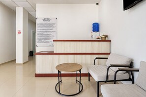 Un área de recepción con un mostrador de madera, una mesa redonda y dos sillas. Hay un cartel informativo grande en la pared y un dispensador de agua sobre el mostrador.