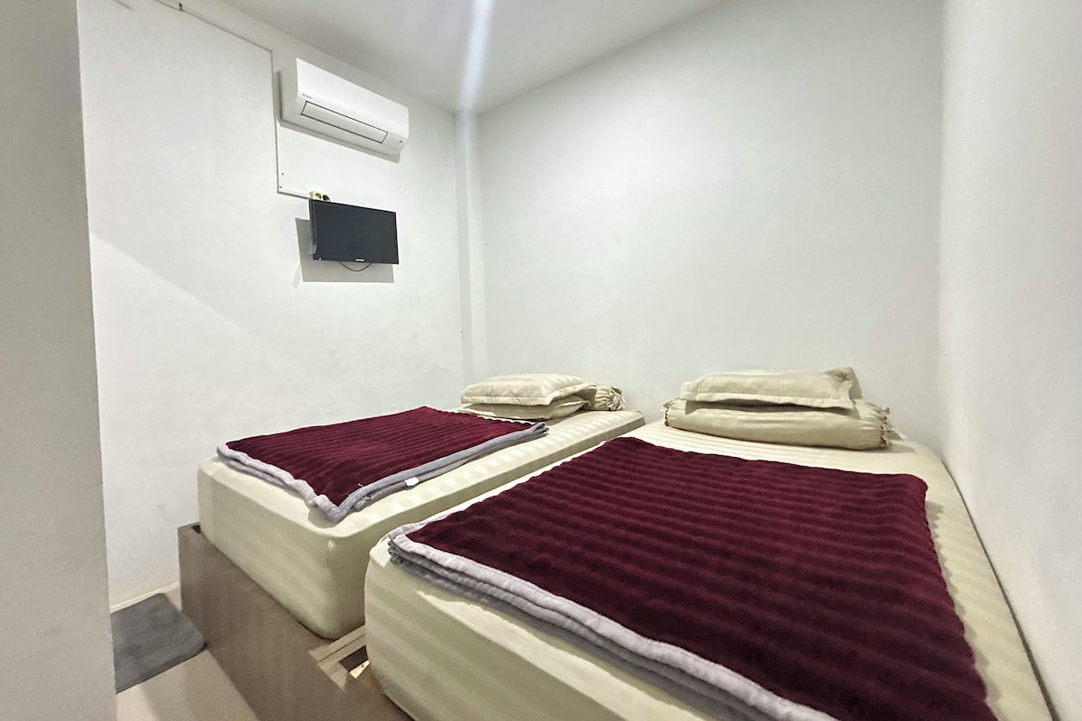 A small, single-room hotel room with two beds, a TV, and an air conditioning unit.