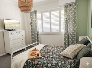 Ein Schlafzimmer mit einem Bett, einem Schminktisch, einem Fernseher, einem Fenster mit Vorhängen und einem Tablett mit Speisen.