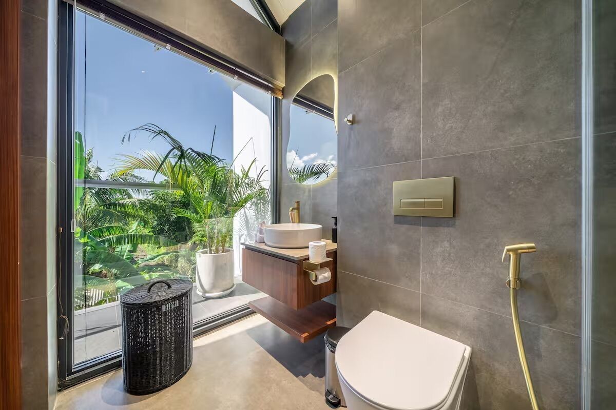 A modern bathroom with a large window, a white toilet, a sink with a mirror, and a shower area.
