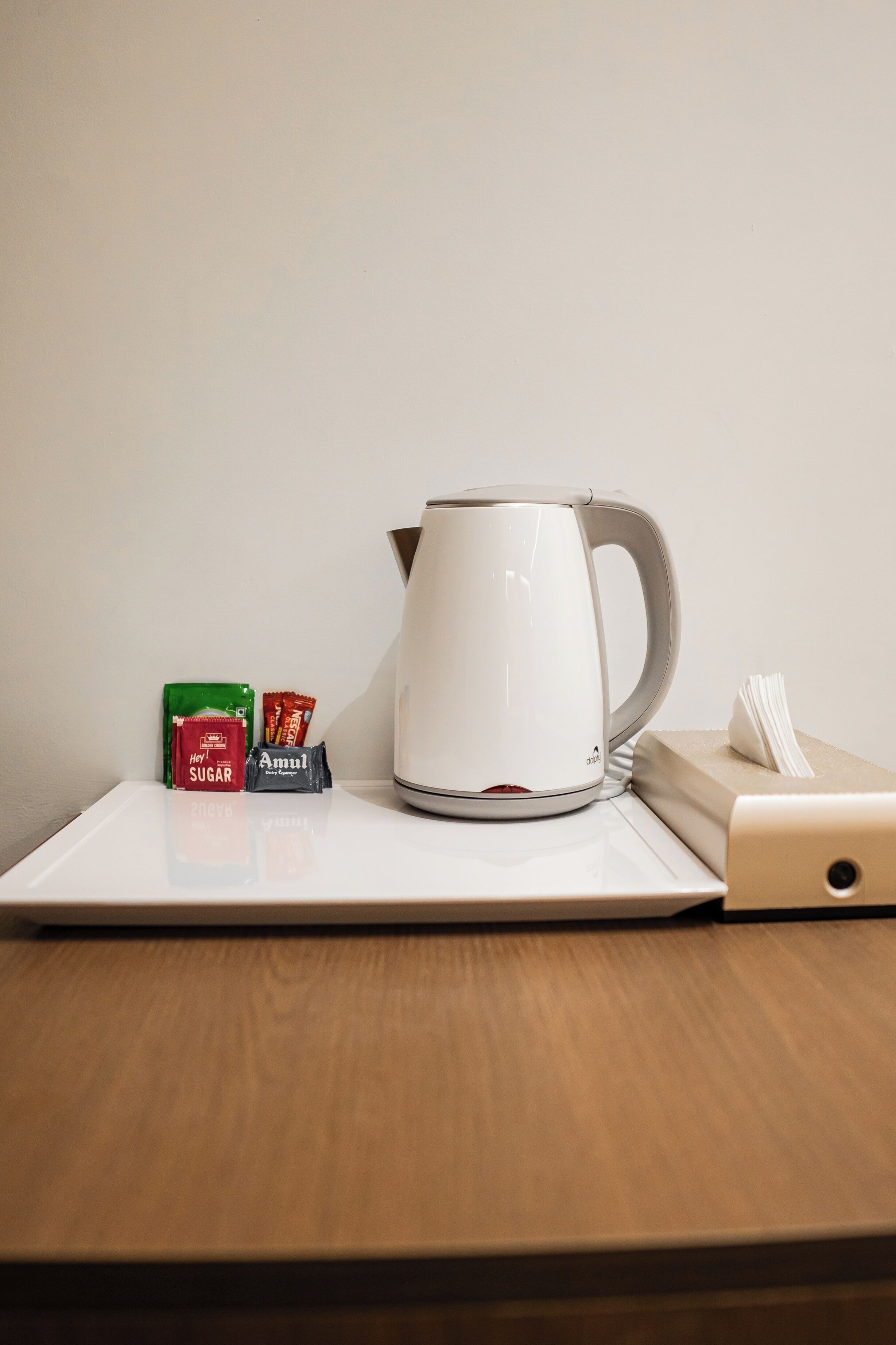 Een wit elektrisch waterkoker op een witte dienblad, een groene doos met de tekst 'Suiker', een rood zakje met 'Amul' en een zakdoos op een houten tafel.