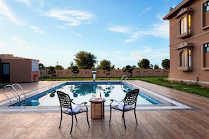 A pool area with two chairs and a table, surrounded by a building with a tiled roof. - ELIVAAS Nirvana 5BHK Farmhouse with Pool (Amer)