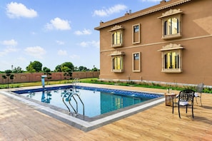 A swimming pool with a wooden deck, a building with ornate windows, and two chairs. - ELIVAAS Nirvana 5BHK Farmhouse with Pool (Amer)