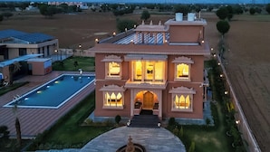 A large, multi-story building with a swimming pool, a fountain, and a well-maintained garden. - ELIVAAS Nirvana 5BHK Farmhouse with Pool (Amer)