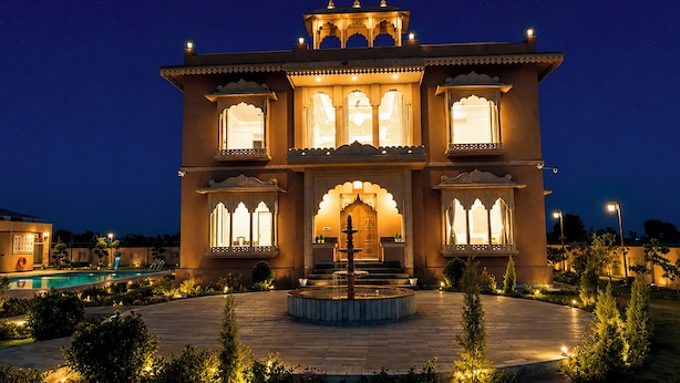 A traditional building with multiple windows, a central entrance, and a fountain in the courtyard. - ELIVAAS Nirvana 5BHK Farmhouse with Pool (Amer)