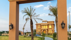 A pathway leading to a building with a traditional architectural style, flanked by palm trees and lamp posts. - ELIVAAS Nirvana 5BHK Farmhouse with Pool (Amer)