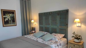 Een bed met een gewatteerde spreideken, twee kussens, een nachtkastje met een lamp, een ingelijst kunstwerk aan de muur en een potplant op een bijzettafeltje.