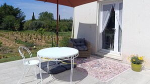 Een terras met een tafel en stoelen, een grote parasol, een rieten bank, een potplant en uitzicht op een wijngaard.