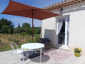 A patio with a table and chairs, a large umbrella, a wicker sofa, a potted plant, and a view of a vineyard. - Charming air-conditioned villa in the vineyards, close to the beach (Roquefort-la-Bédoule)