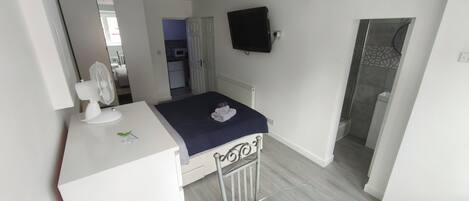 Et soveværelse med en seng, et skrivebord med ventilator, et fjernsyn på væggen og et badeværelse, der kan ses gennem en åben dør.