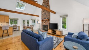 Uma sala de estar espaçosa com teto alto, vigas de madeira aparentes, lareira de tijolos, televisão e área de jantar com mesa e cadeiras.