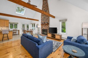 A spacious living area with a high ceiling, exposed wooden beams, a brick fireplace, a television, and a dining area with a table and chairs. - Walk to Big Sandy Pond Cape Escape (West Yarmouth)