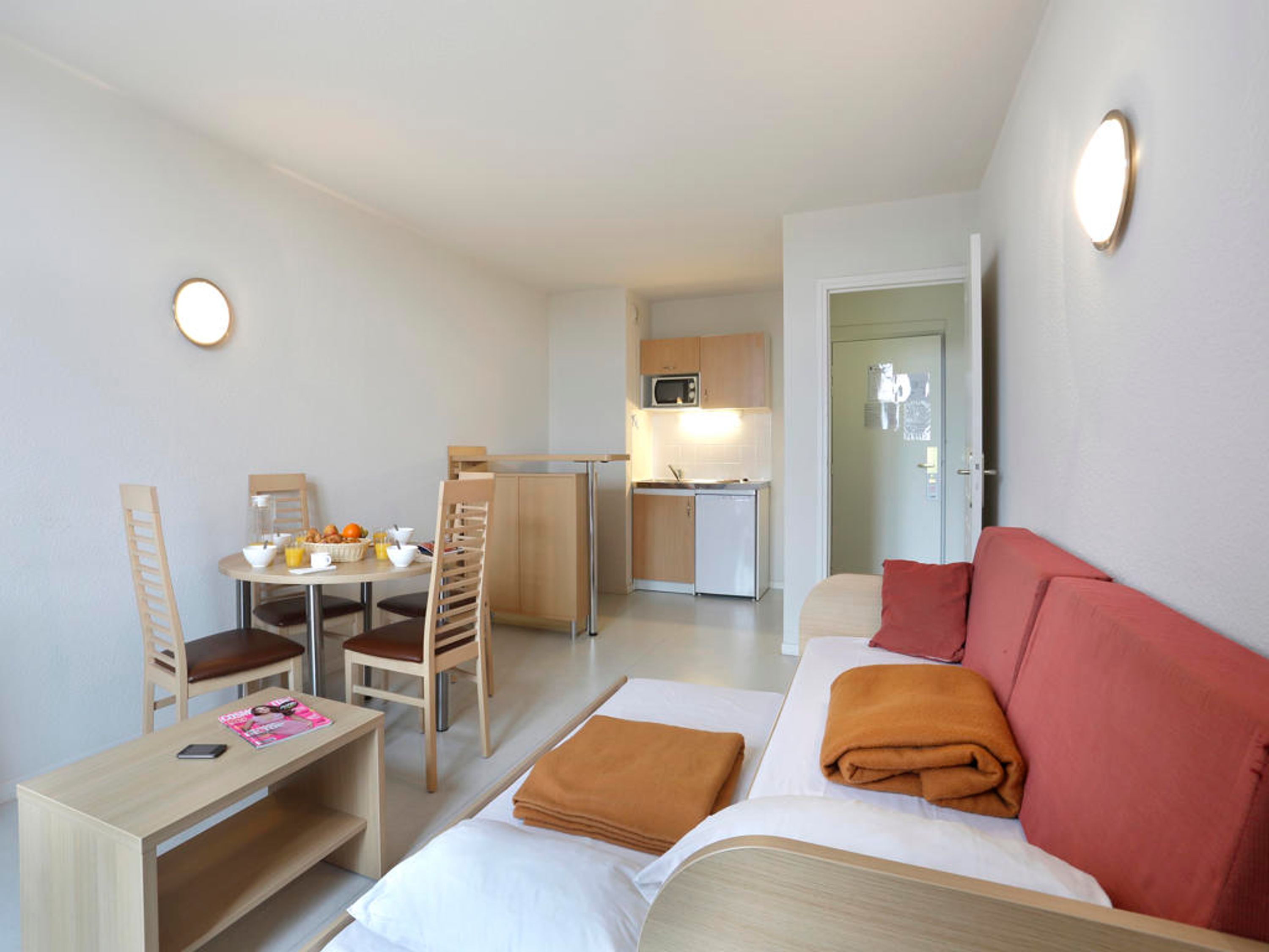 A compact hotel room with a kitchenette, a dining area, and a bed with a red accent pillow.