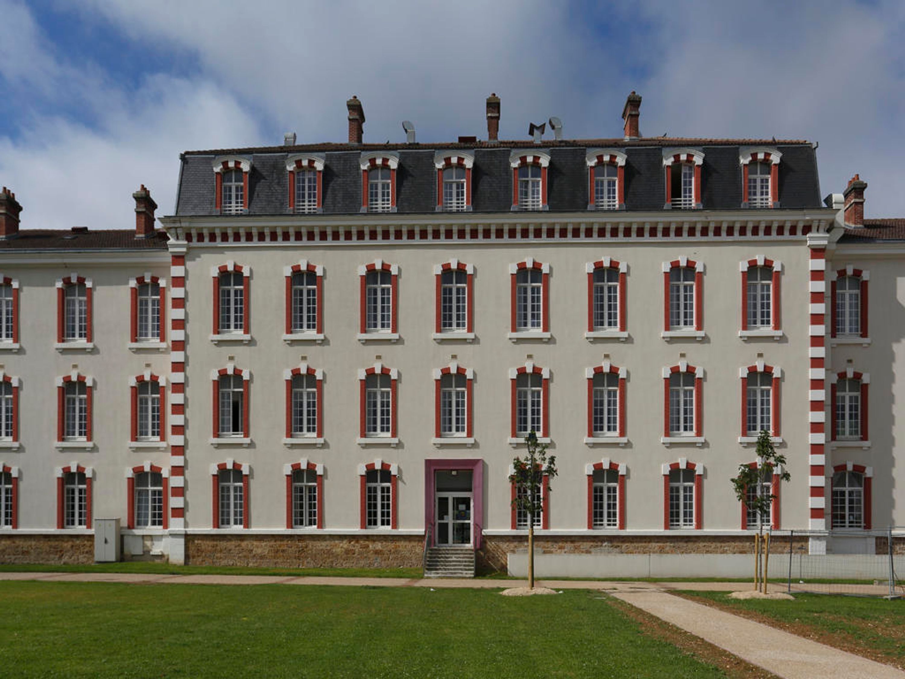 A large, symmetrical building with a central entrance, multiple windows with red shutters, and a flat roof with chimneys.