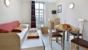 Una habitación de hotel pequeña con cama, un sofá compacto, una mesa de comedor con sillas, televisión y ventana con cortinas.