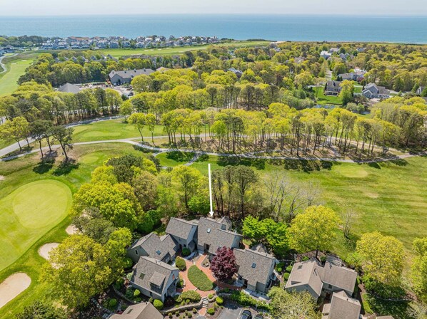 An aerial view of a residential area with houses, a golf course, and a flagpole. - Sea Spark - Cape Escape with Pool Access (Mashpee)