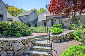 A residential area with stone steps leading to a house, surrounded by well-maintained hedges and a variety of trees. - Sea Spark - Cape Escape with Pool Access (Mashpee)