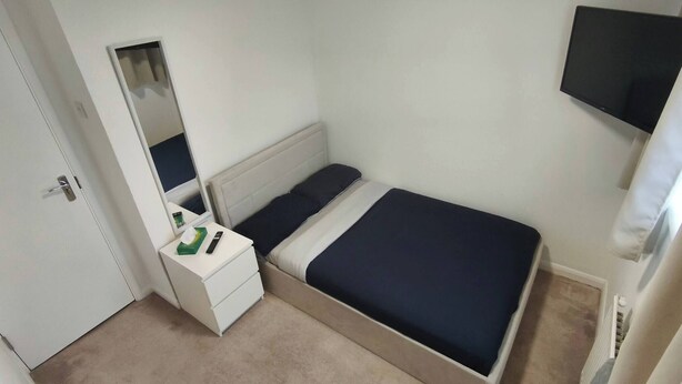 A small, single-bed room with a white bedside table, a mirror, a flat-screen TV mounted on the wall, and a door. - Room-small Double in Quiet House - Chelm (Chelmsford)