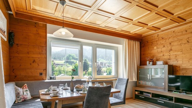 A cozy room with a wooden ceiling, a dining table with chairs, a sofa, a television, and a window with a view of greenery. - Dreamlike vacation apartment with sunny terrace. (Wildschönau)