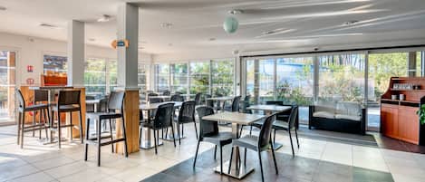 A spacious indoor dining area with multiple tables and chairs, large windows offering a view of the outdoors, and a seating area with a sofa and a cabinet.
