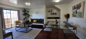 A modern living room with a glass coffee table, a sofa with yellow cushions, a dining table with chairs, and a large plant.
