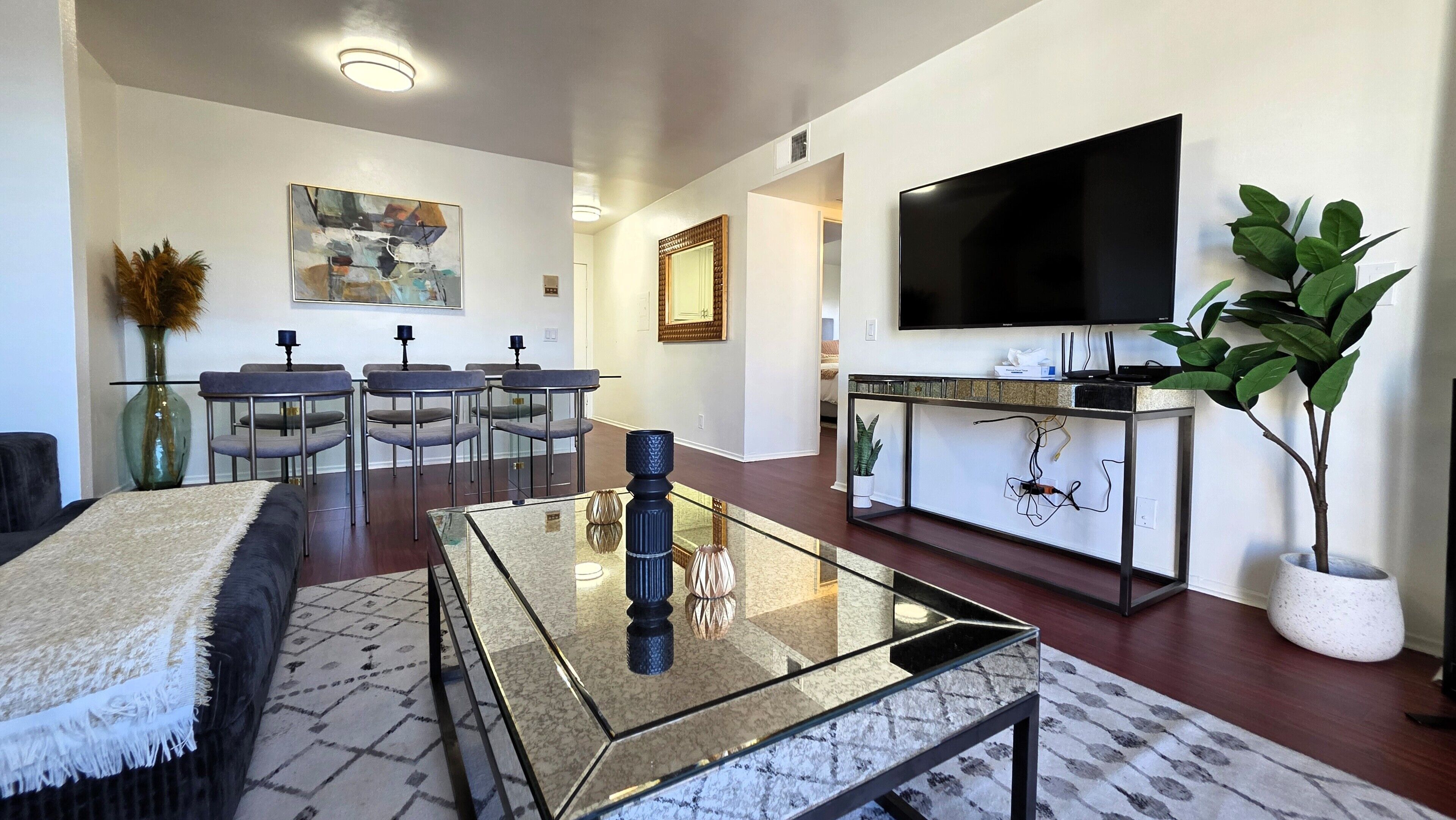 A modern living room with a glass coffee table, a flat-screen TV, a dining area with chairs, and a potted plant.
