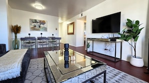 A modern living room with a glass coffee table, a flat-screen TV, a dining area with chairs, and a potted plant.