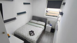 A small, neatly arranged bedroom with a bed, bedside table, and a window with blinds. - Room-cozy Double Room Near City Centre & (Chelmsford)
