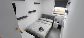 A small, neatly arranged bedroom with a bed, bedside table, and a window with blinds.
