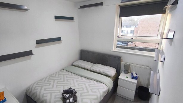 A bedroom with a bed, bedside table, and a window with a view of buildings. - Room-cozy Double Room Near City Centre & (Chelmsford)