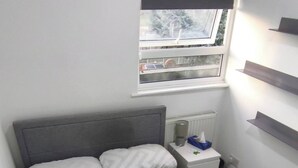 A bed with a patterned bedspread, a bedside table with a tissue box, a window with a view, and built-in shelves. - Room-cozy Double Room Near City Centre & (Chelmsford)